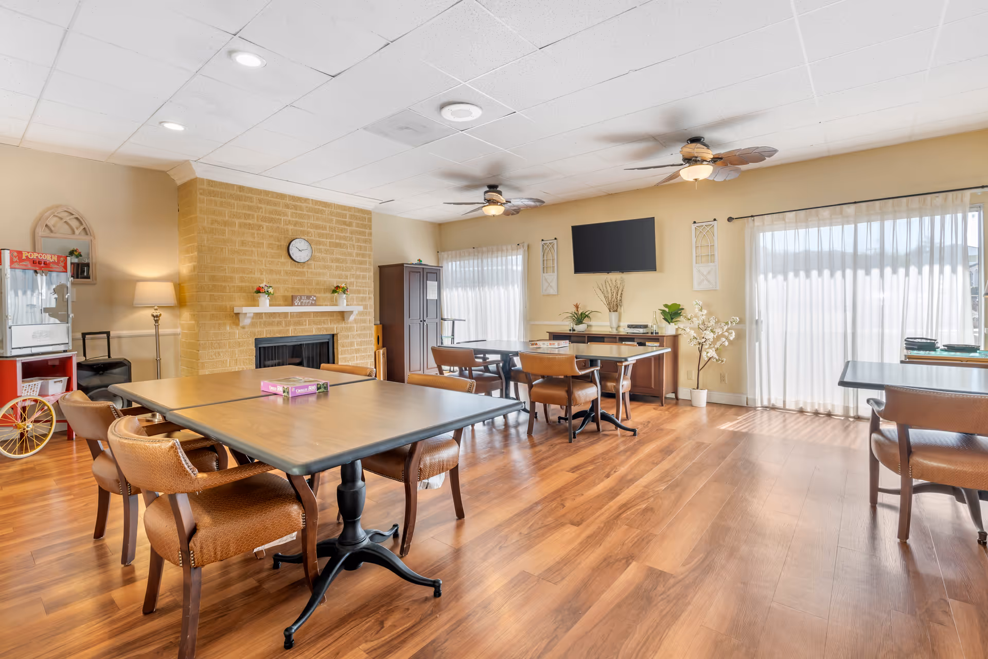 Spacious senior living common area with tables, chairs, a fireplace, and large windows.
