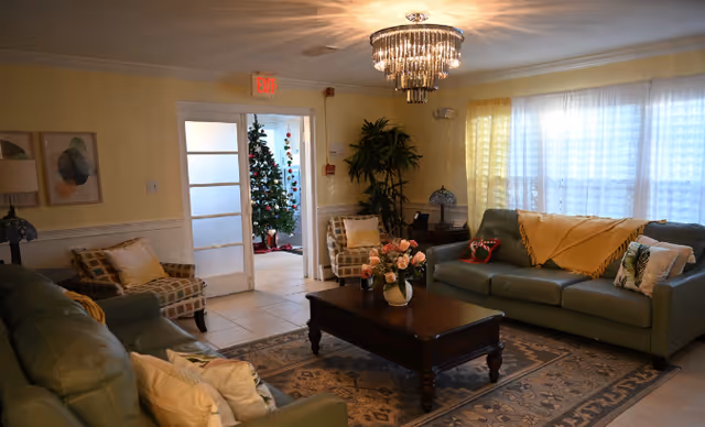 Cozy senior living room with green sofas, a coffee table, plants, chandelier, and doorway.