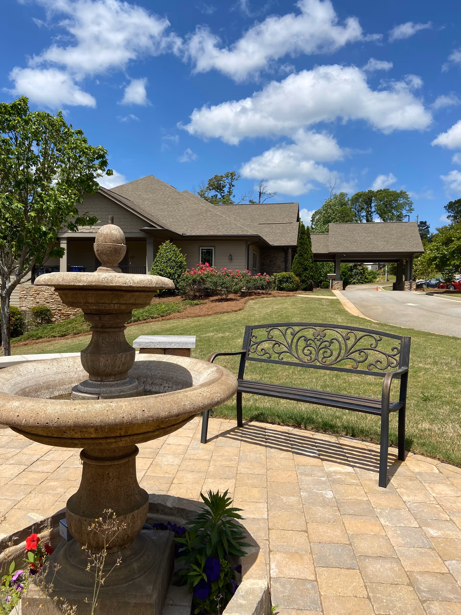 A stone fountain and bench on a sunny patio in front of a beige hospice building.