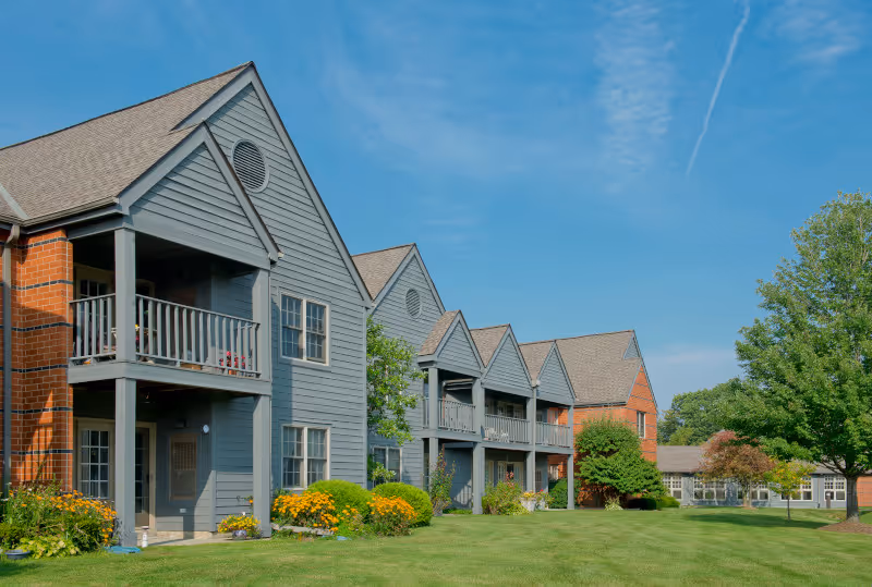 Exterior view of a senior living facility with multiple two-story buildings featuring gray siding and brick accents, balconies, and well-maintained green lawns with trees and shrubs under a clear blue sky.