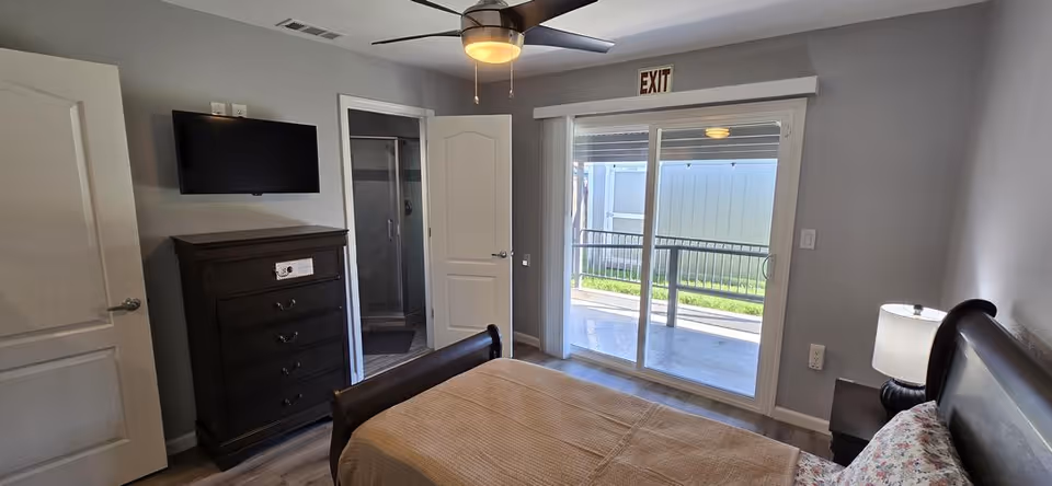 A furnished bedroom in a senior living facility with bed, dresser, TV, and balcony.