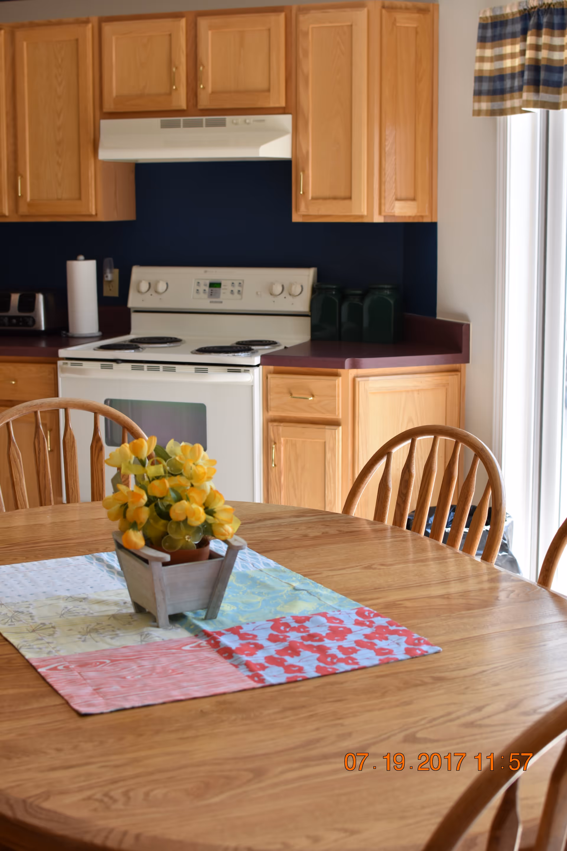 A kitchen area with wooden cabinets, a white electric stove with an overhead vent, and a countertop with three green canisters. In the foreground, there is a wooden dining table with a colorful patchwork table runner and a small potted plant with yellow flowers. A window with a blue and beige plaid valance is visible on the right side.