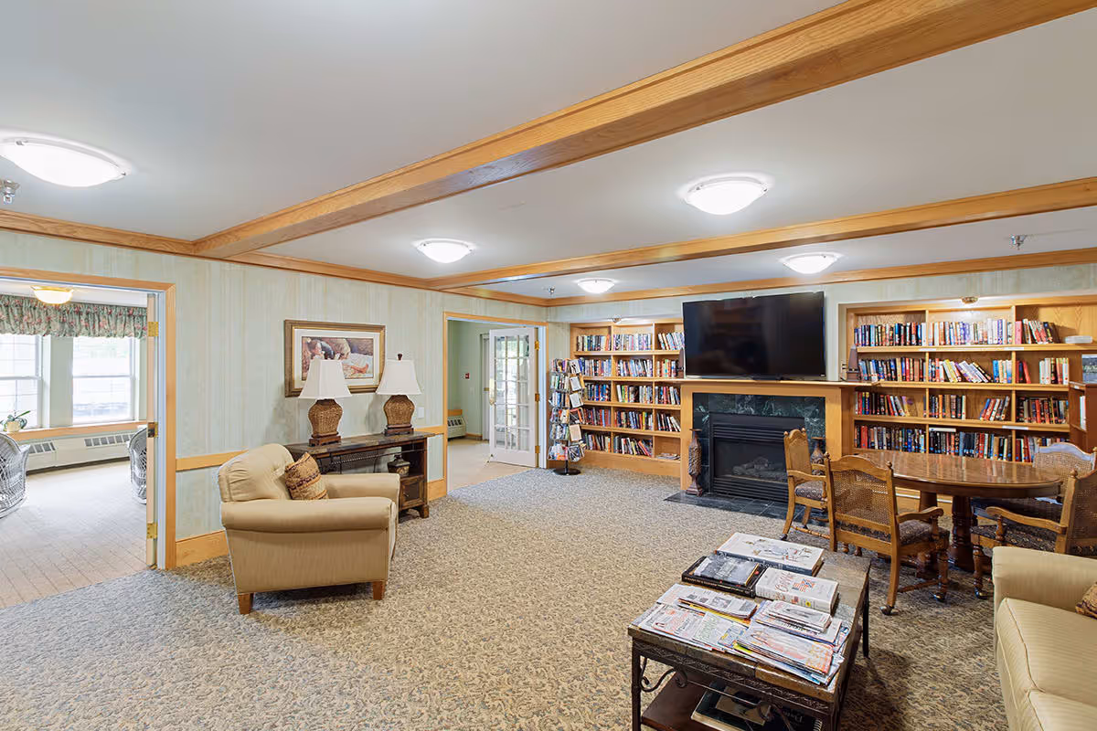 A cozy living room area in Eagle Crest Assisted Living featuring beige armchairs, a coffee table with newspapers, a round wooden table with chairs, built-in bookshelves filled with books, a fireplace with a mounted flat-screen TV above it, and soft ceiling lighting. The room has light-colored walls with wooden trim and a carpeted floor.