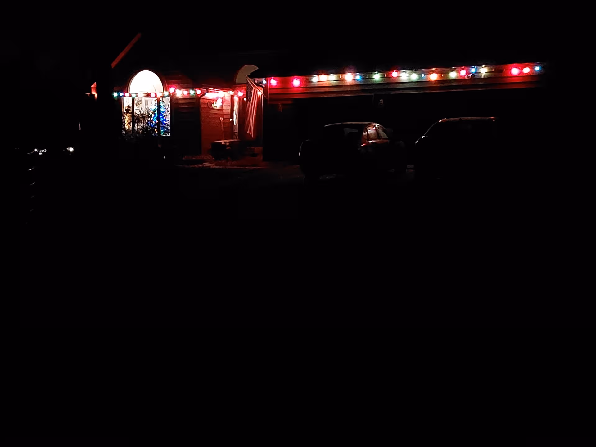 Nighttime exterior of a building entrance decorated with colorful string lights and parked cars.