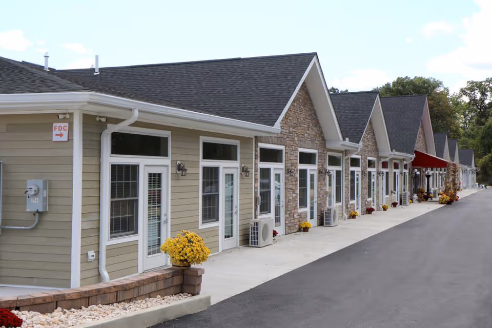 Exterior view of a single-story building with multiple entrances, each featuring white-framed glass doors and windows. The building has a combination of beige siding and stone facade, with a dark shingled roof. Yellow potted flowers are placed along the walkway, and there is a red awning further down the building. The area is paved with asphalt and concrete walkways, with trees visible in the background under a partly cloudy sky.