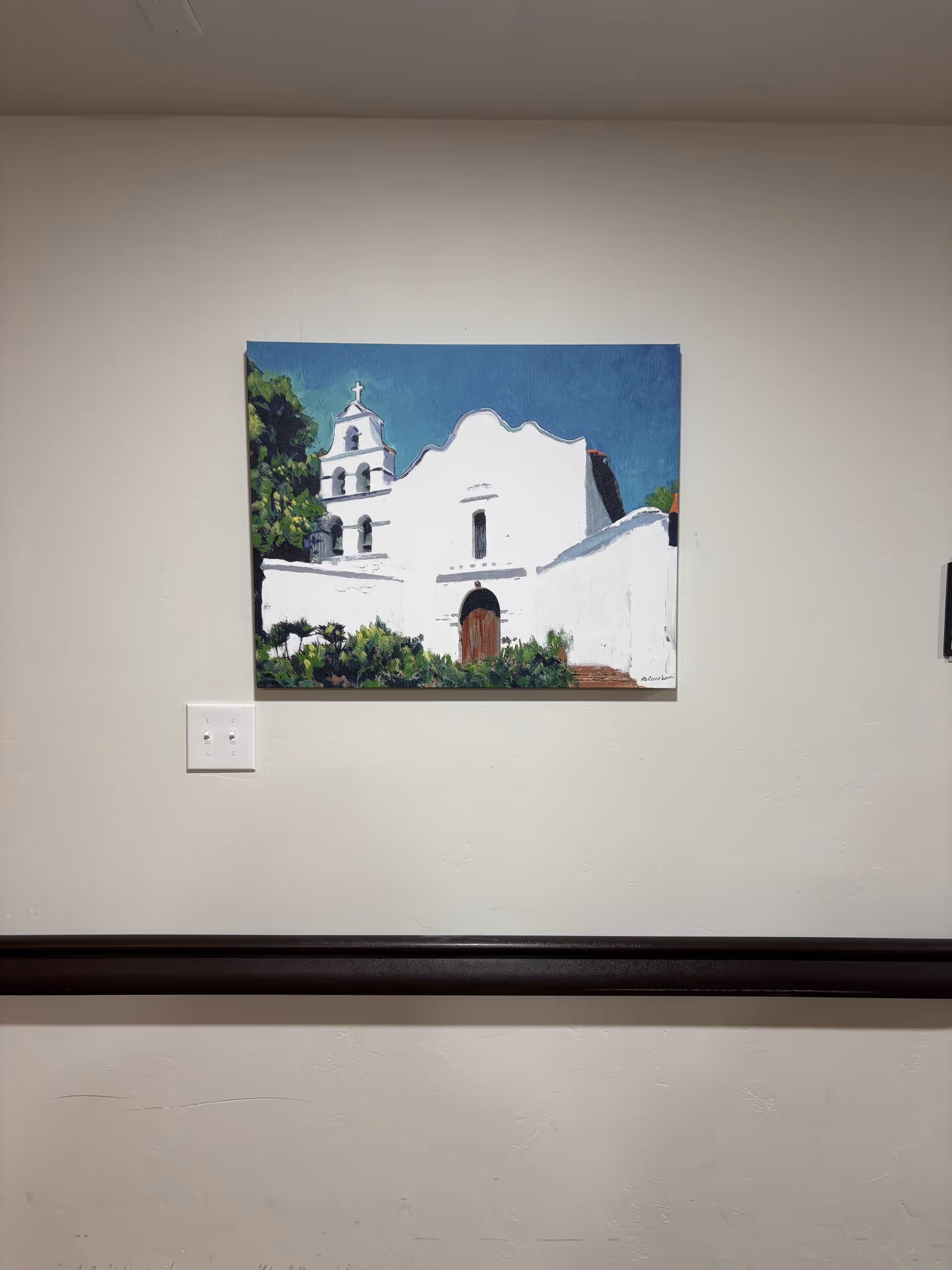 Interior hallway wall featuring a painting of a white building and a black railing.