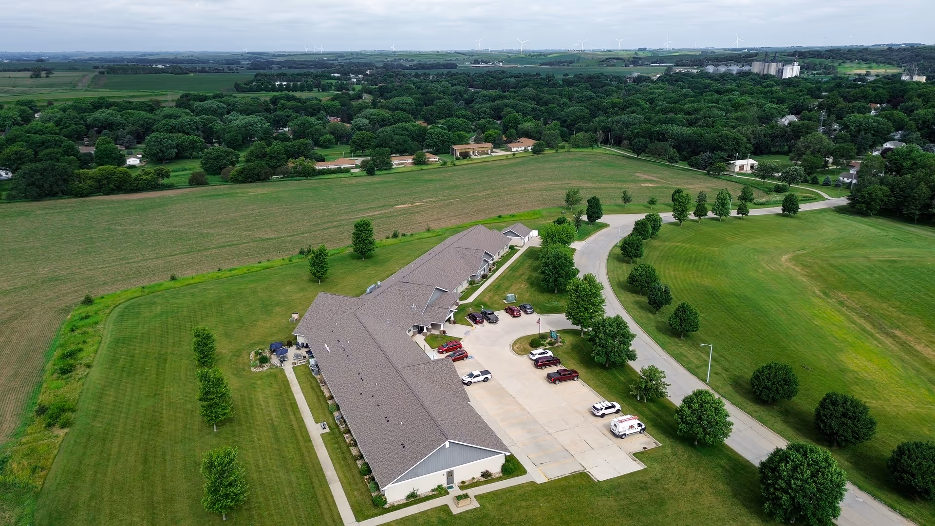 Aerial view of a single-story senior living facility named Golden Horizons of Ida Grove surrounded by green lawns and trees, with a parking lot containing several vehicles and a curved driveway leading to the building.