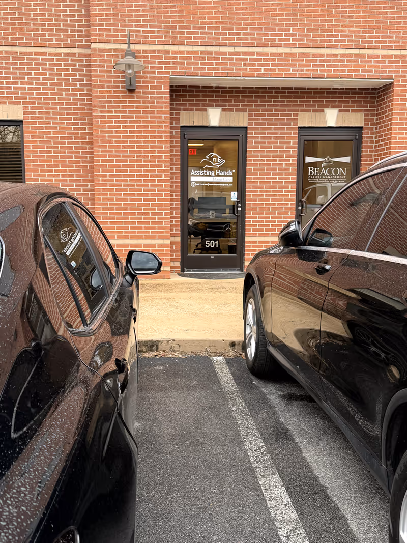 Brick building entrance with glass doors bearing Assisting Hands and Beacon logos; parked cars in foreground.