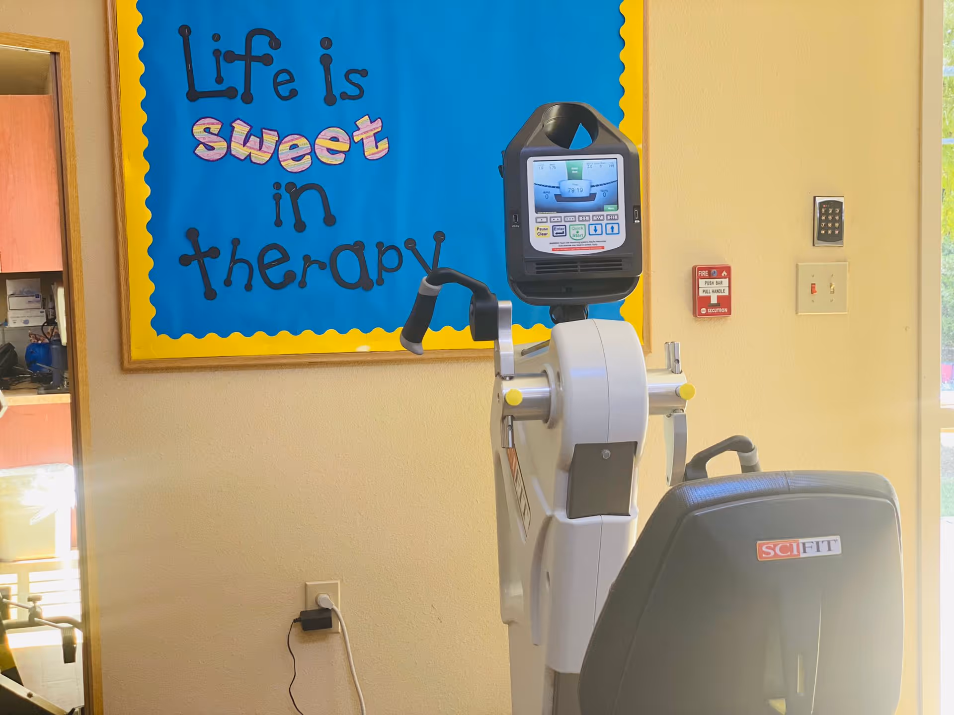 A therapy exercise machine with a digital display and handles is positioned in front of a wall with a colorful bulletin board that reads 'Life is sweet in therapy'. There is a fire alarm and a keypad on the wall, and a window letting in natural light.