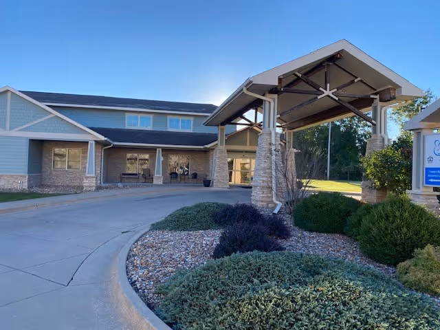 Entrance of a two-story senior living building with a covered porte-cochere, driveway, and landscaped rock and shrub beds.
