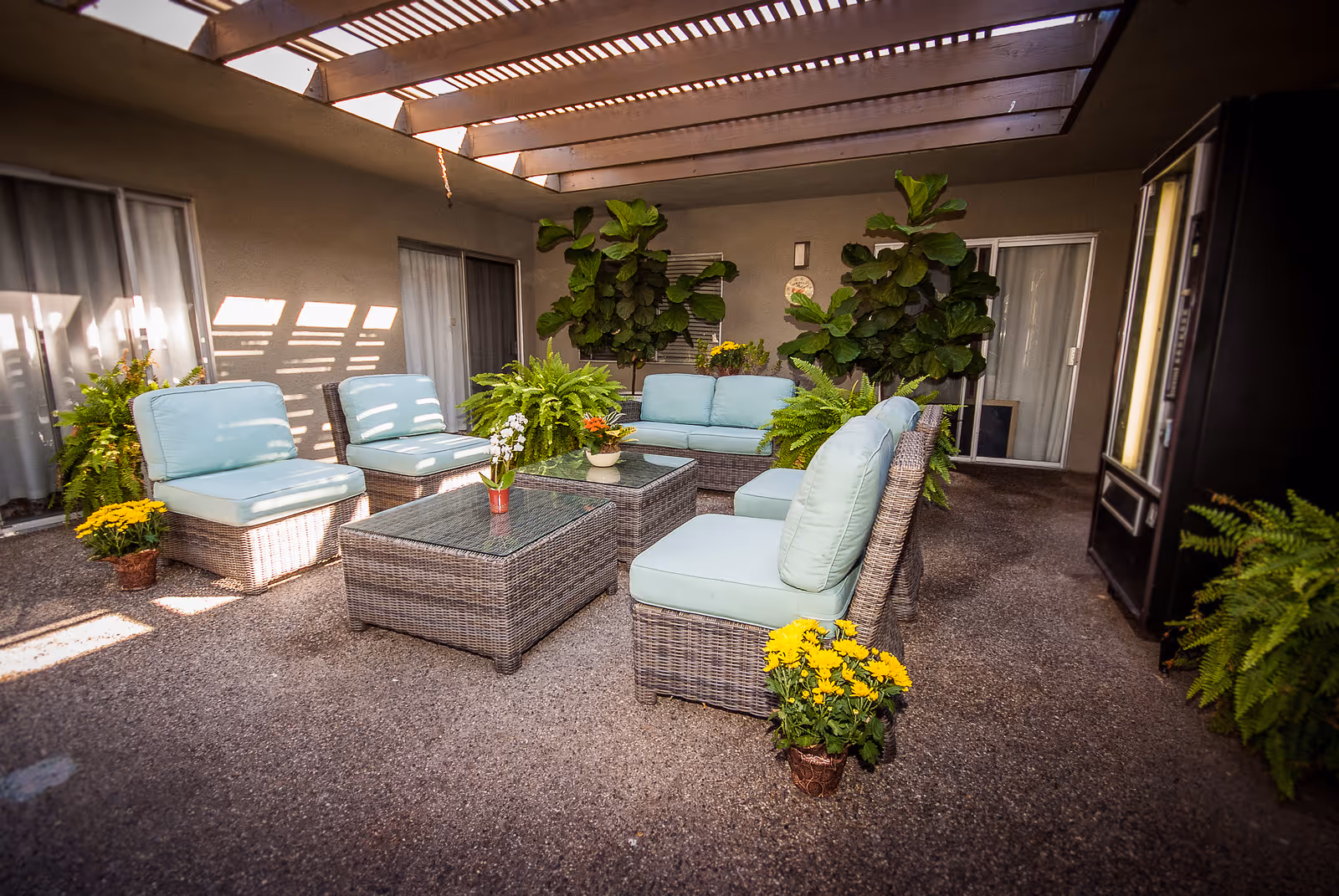 Outdoor covered patio area with wicker furniture including chairs and a sofa with light blue cushions, glass-top coffee tables, and various potted plants and flowers. The area has a pergola-style roof allowing sunlight to filter through, and sliding glass doors lead to the interior.