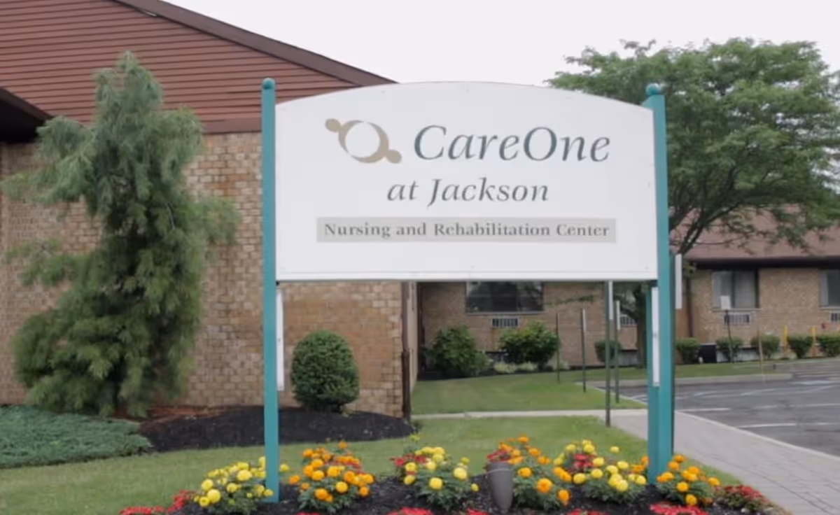 Outdoor view of a sign for CareOne at Jackson Nursing and Rehabilitation Center, surrounded by a flower bed with yellow and orange flowers, with a brick building and trees in the background.