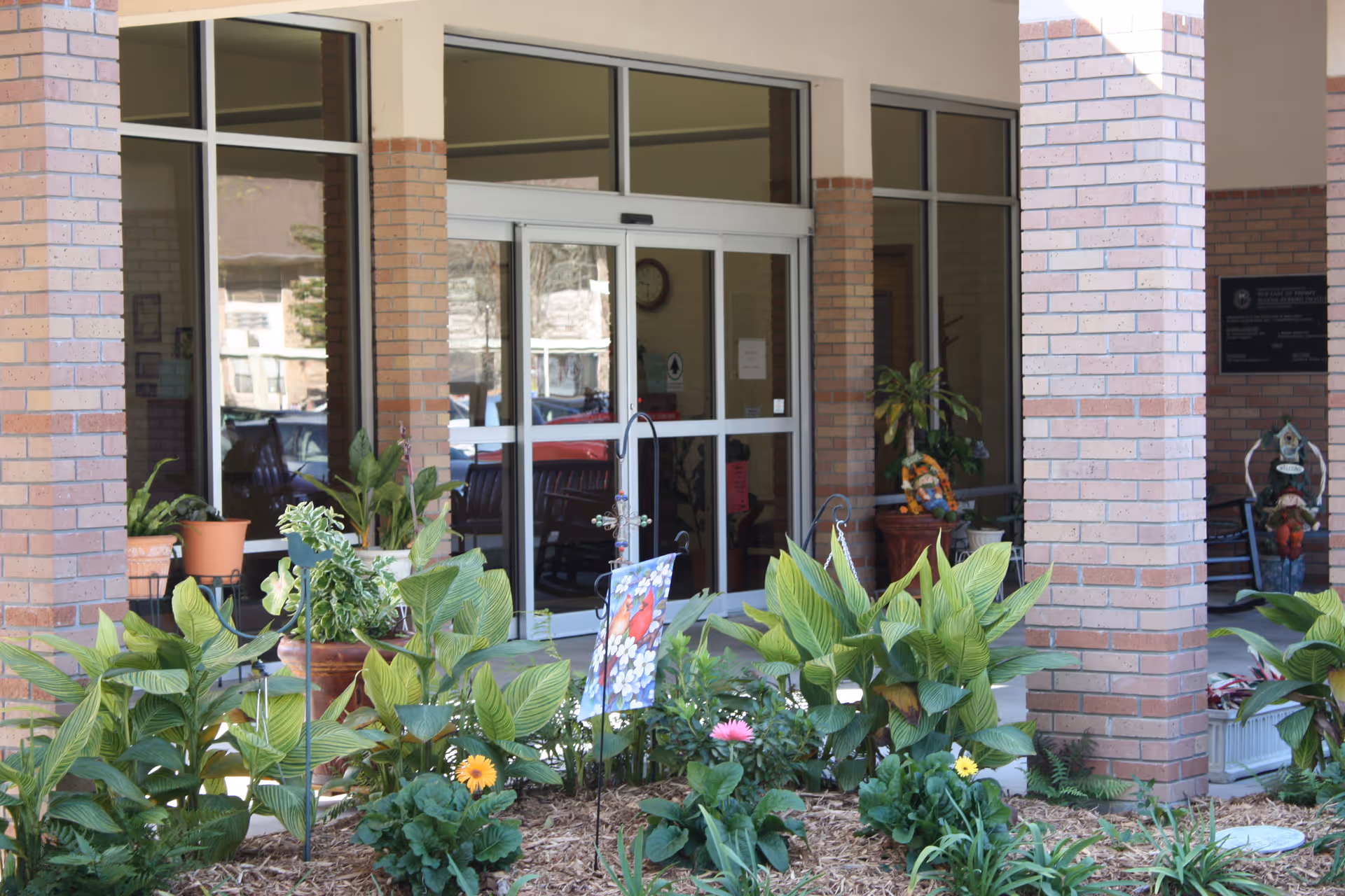 Entrance to a building with large glass doors and windows framed by brick columns. In front of the entrance is a garden bed with various green plants and colorful flowers. There are potted plants on either side of the entrance and a small decorative flag with a bird design in the garden.