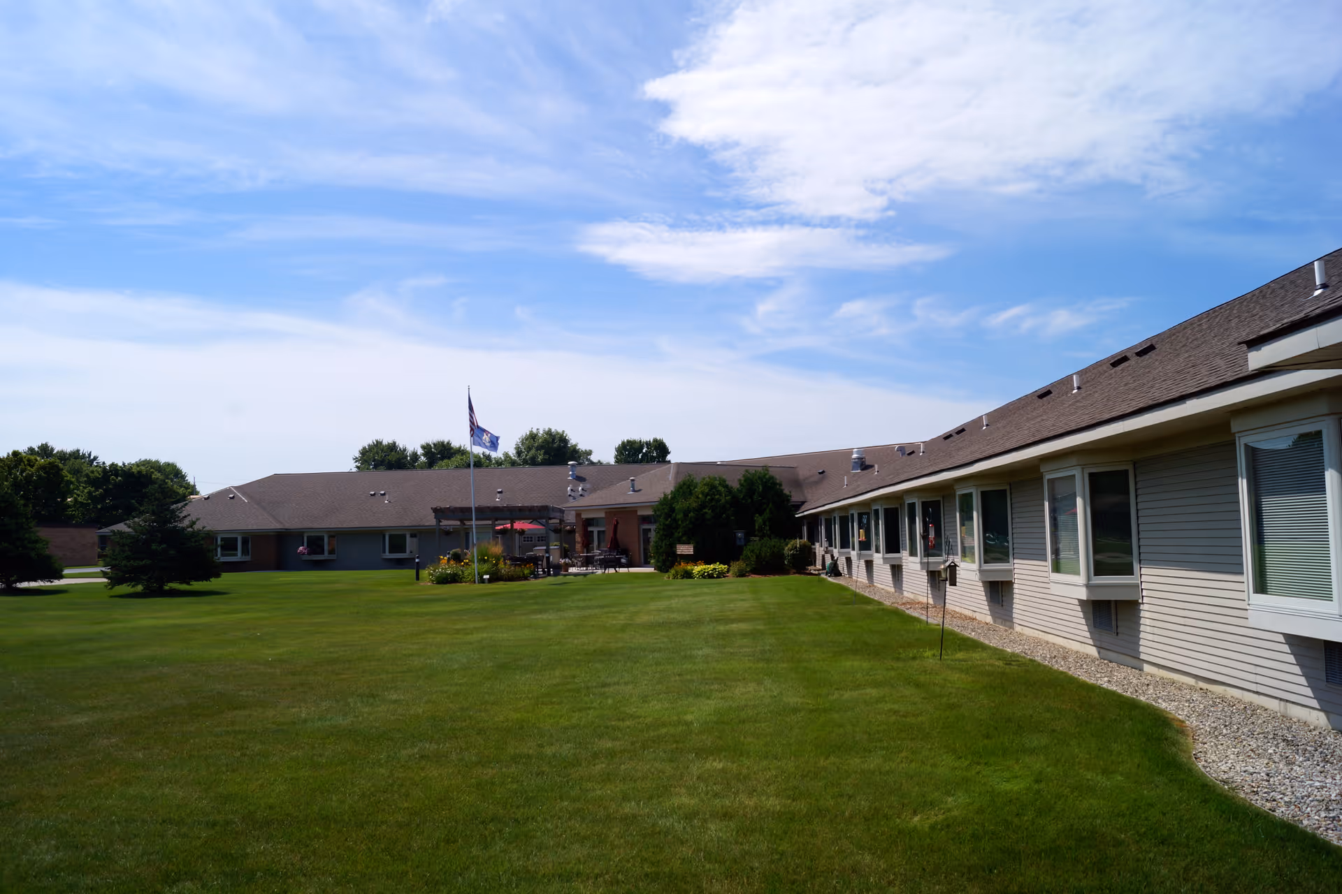 Exterior view of a single-story assisted living facility building with a large green lawn in front, a flagpole with flags, and a partly cloudy blue sky above.