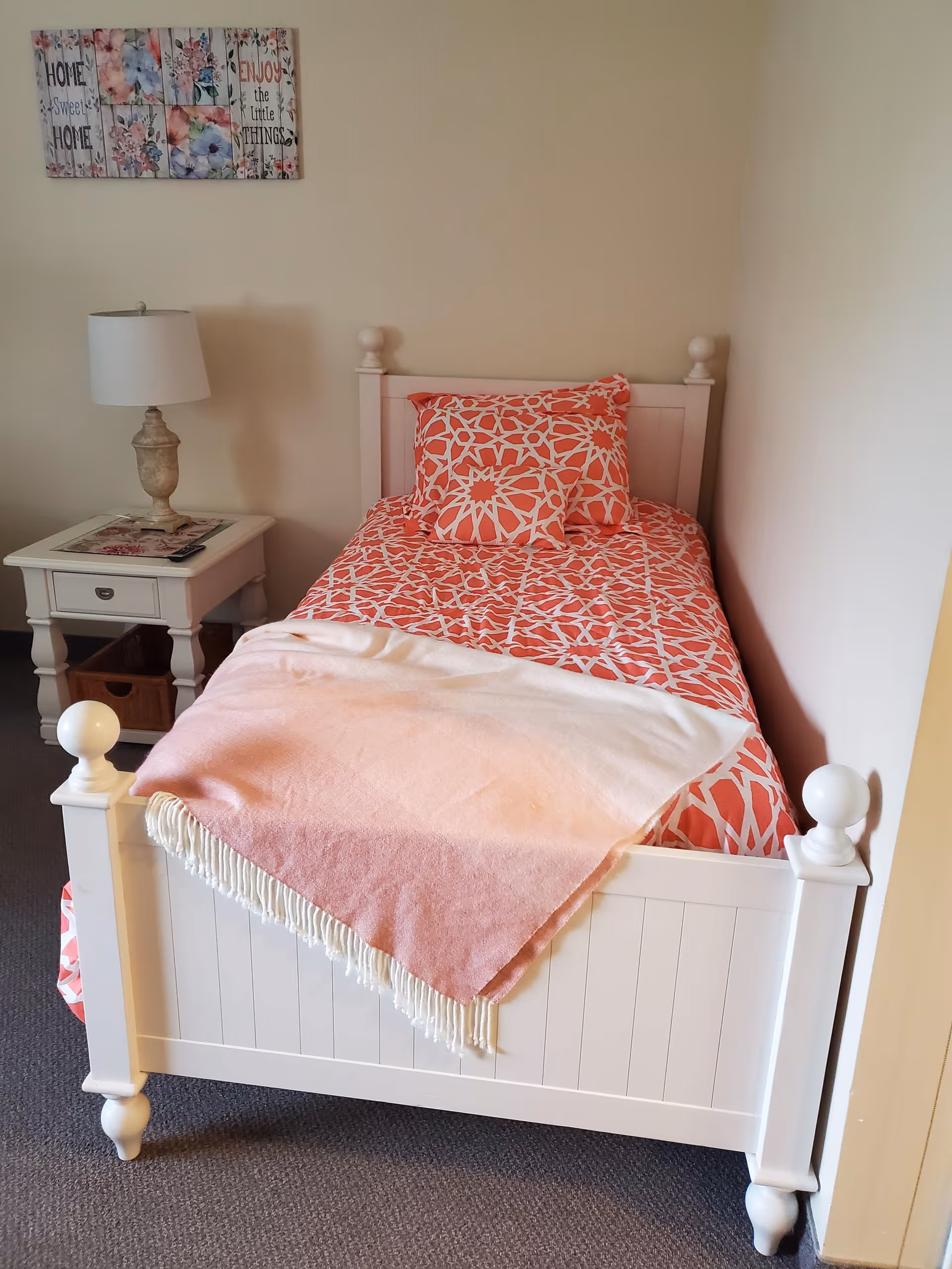 A small bedroom with a white wooden single bed featuring a coral and white patterned bedspread and matching pillows. A pink and white throw blanket is draped over the foot of the bed. Next to the bed is a white nightstand with a lamp and a remote control on top. A decorative wall sign above the nightstand reads 'Home Sweet Home' and 'Enjoy the Little Things'.