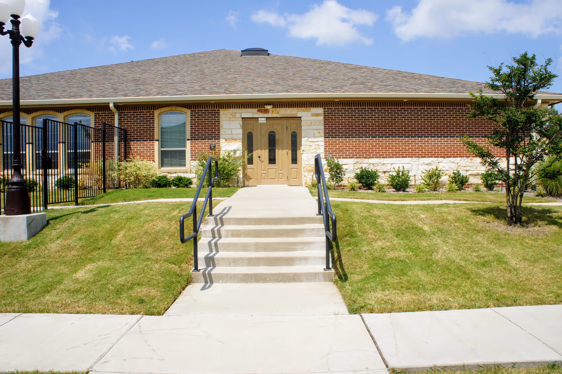 Front exterior view of a single-story brick building with a beige door, steps with black handrails leading up to the entrance, a lamp post on the left, and a small tree on the right under a blue sky with some clouds.