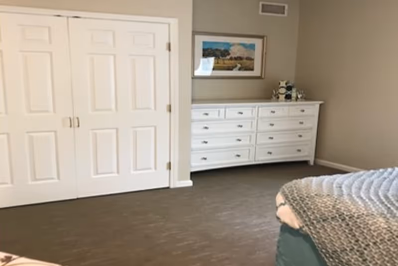 Tidy bedroom featuring a white dresser, framed landscape painting, and a partially visible bed.