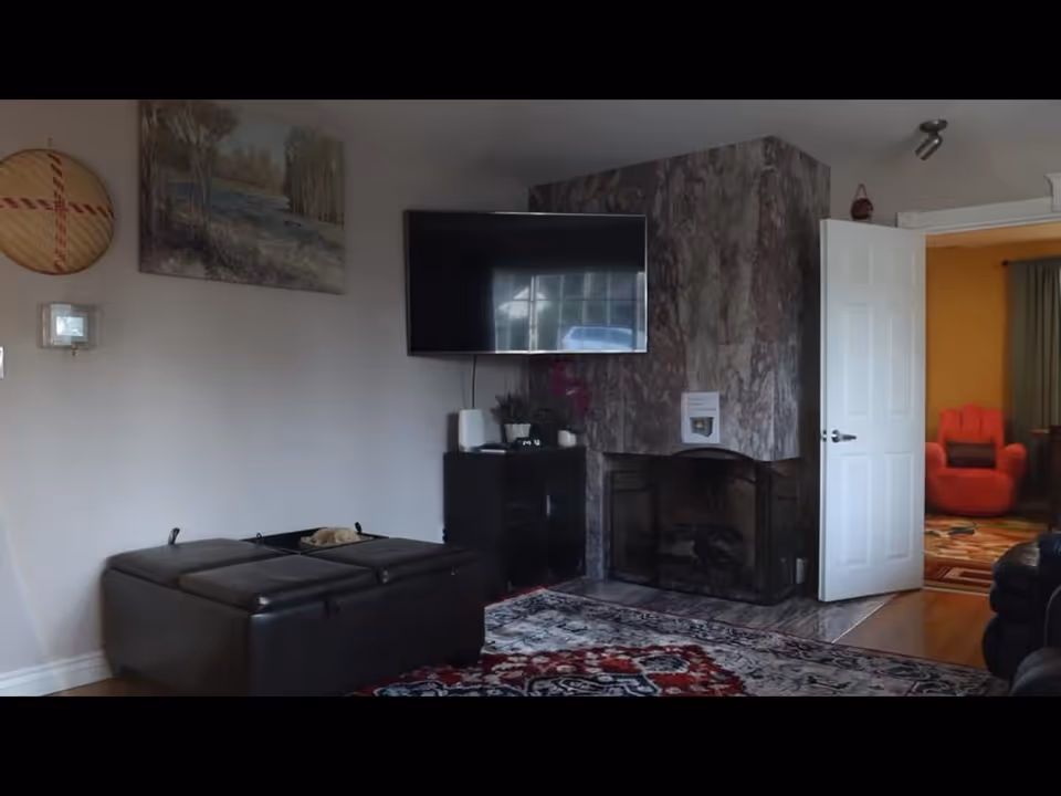 Living room with mounted TV, marble fireplace, dark leather ottomans, and doorway to adjoining room.