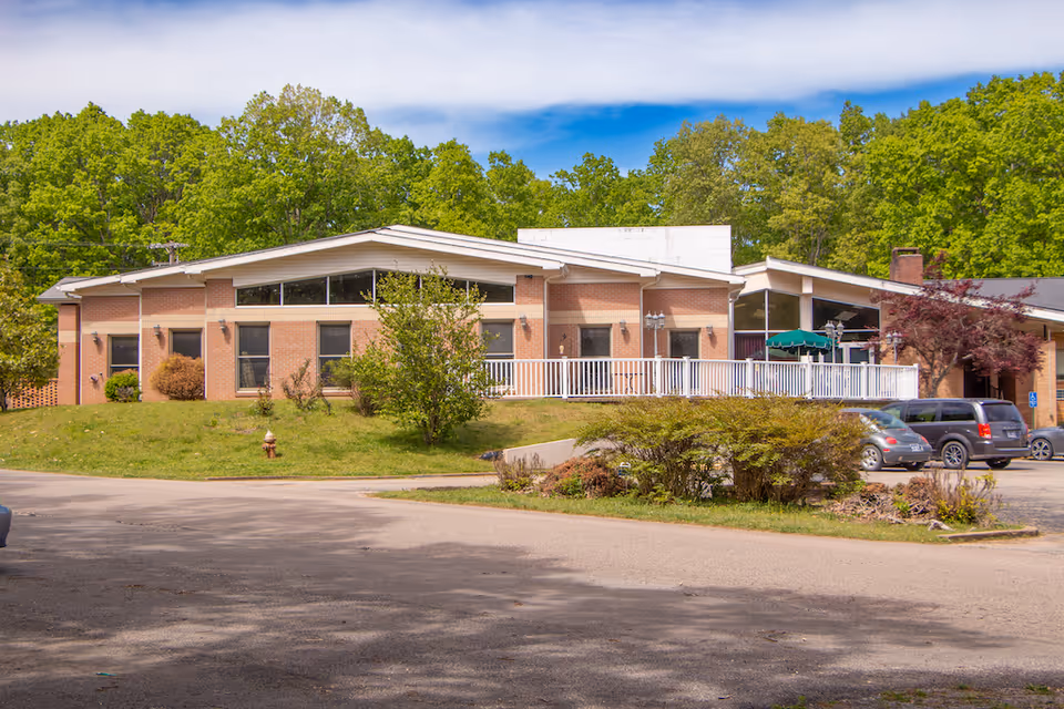 Exterior view of a single-story brick building with large windows and a white railing along a raised patio area. Several cars are parked in front, and the building is surrounded by green trees and shrubs under a partly cloudy sky.