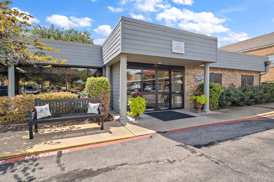 Exterior view of the main entrance to Morada Temple facility featuring a bench with two decorative pillows, potted plants, and a glass door entrance under a gray overhang with a sign that reads 'Main Entrance'.