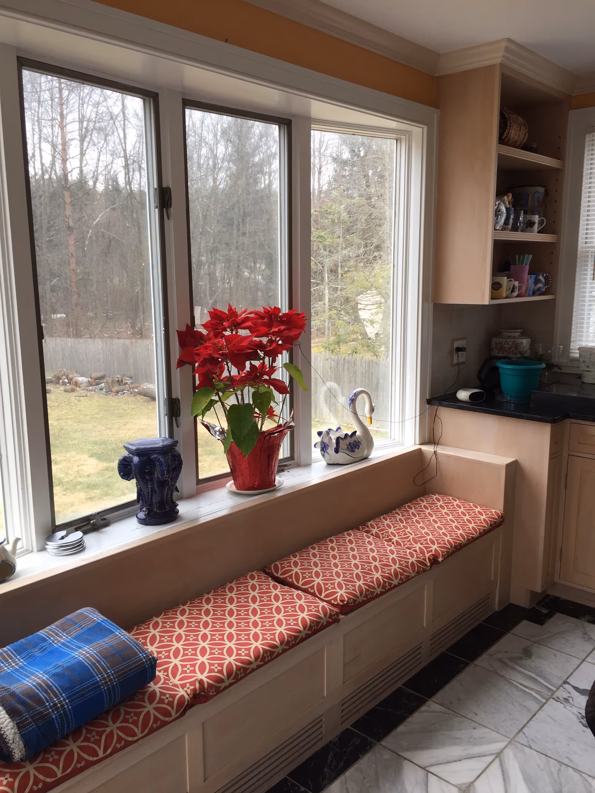 Bright kitchen seating by a bay window with red patterned cushions and mugs on shelves.