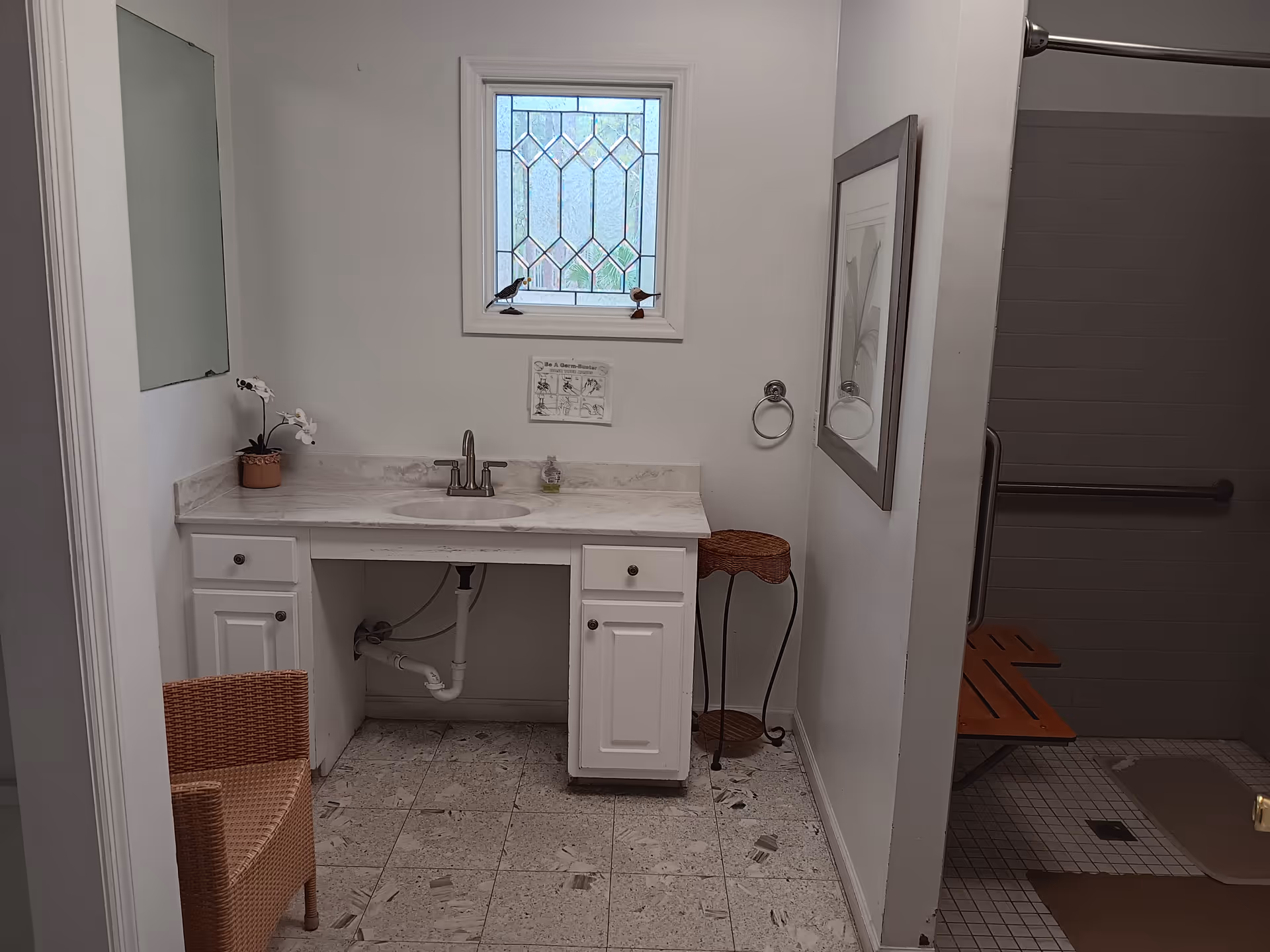 Small bathroom with white vanity, sink, mirror, window, wicker chair, and shower area.