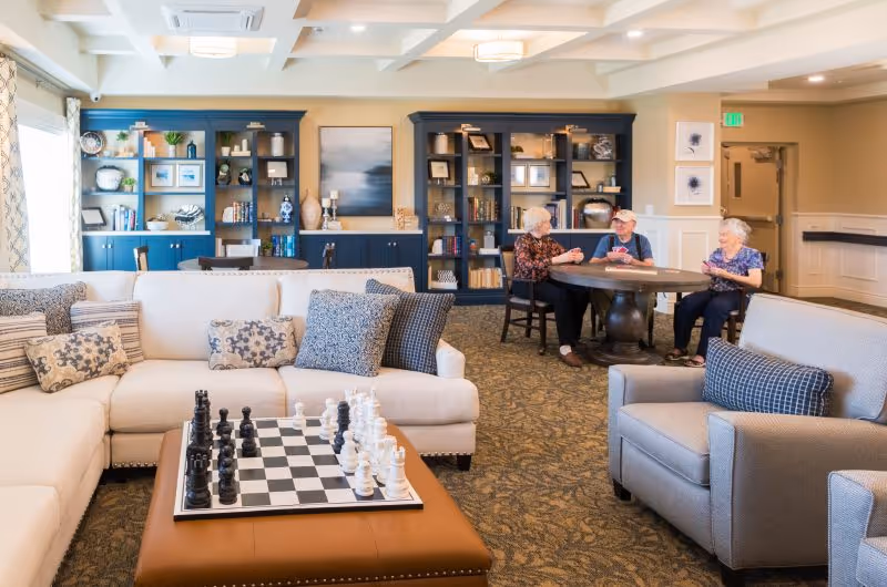 Cozy senior living lounge featuring sofas, a chessboard, shelves, and residents socializing.