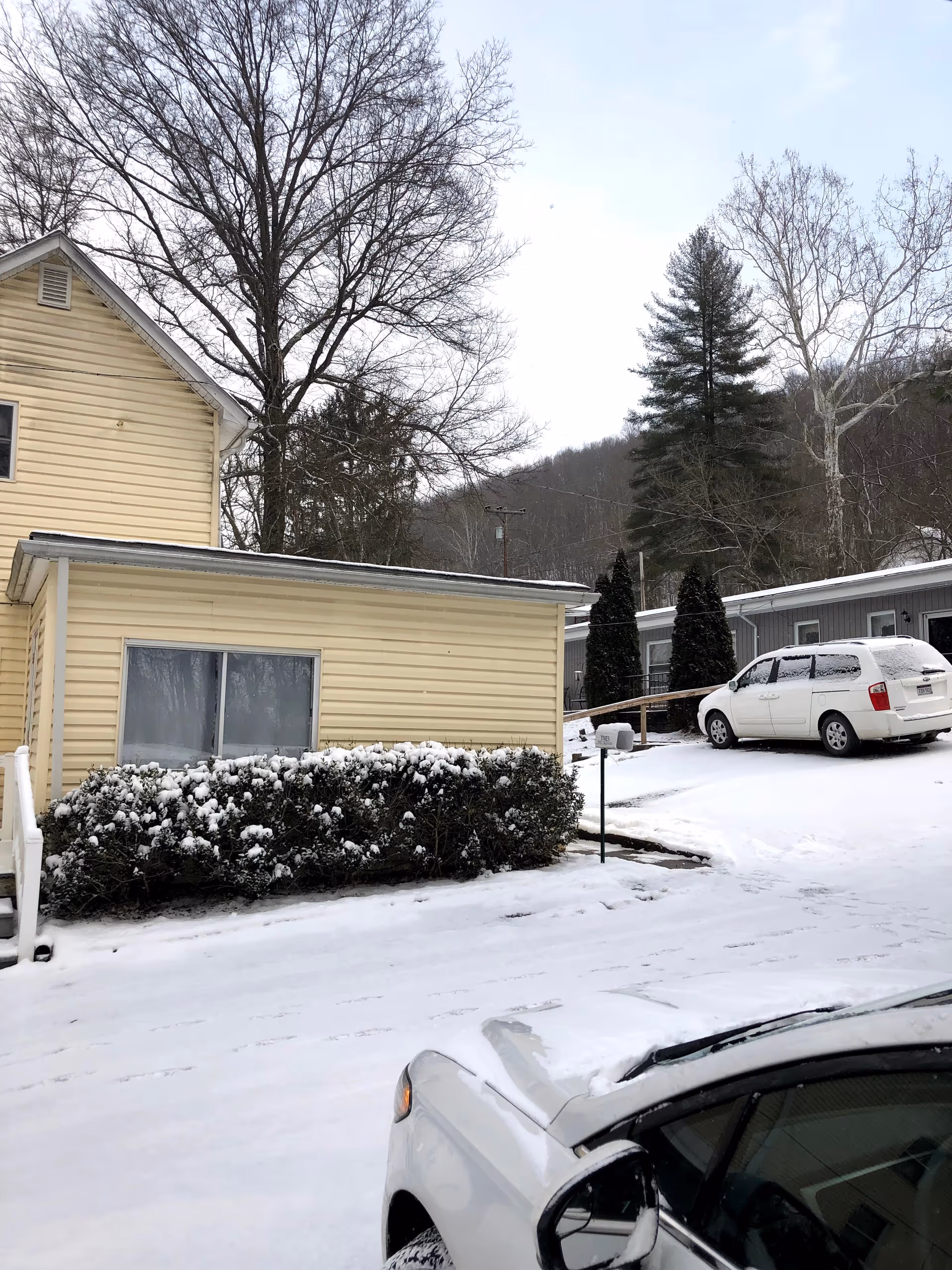 Exterior view of a beige assisted living building with snow, shrubs, and parked cars.