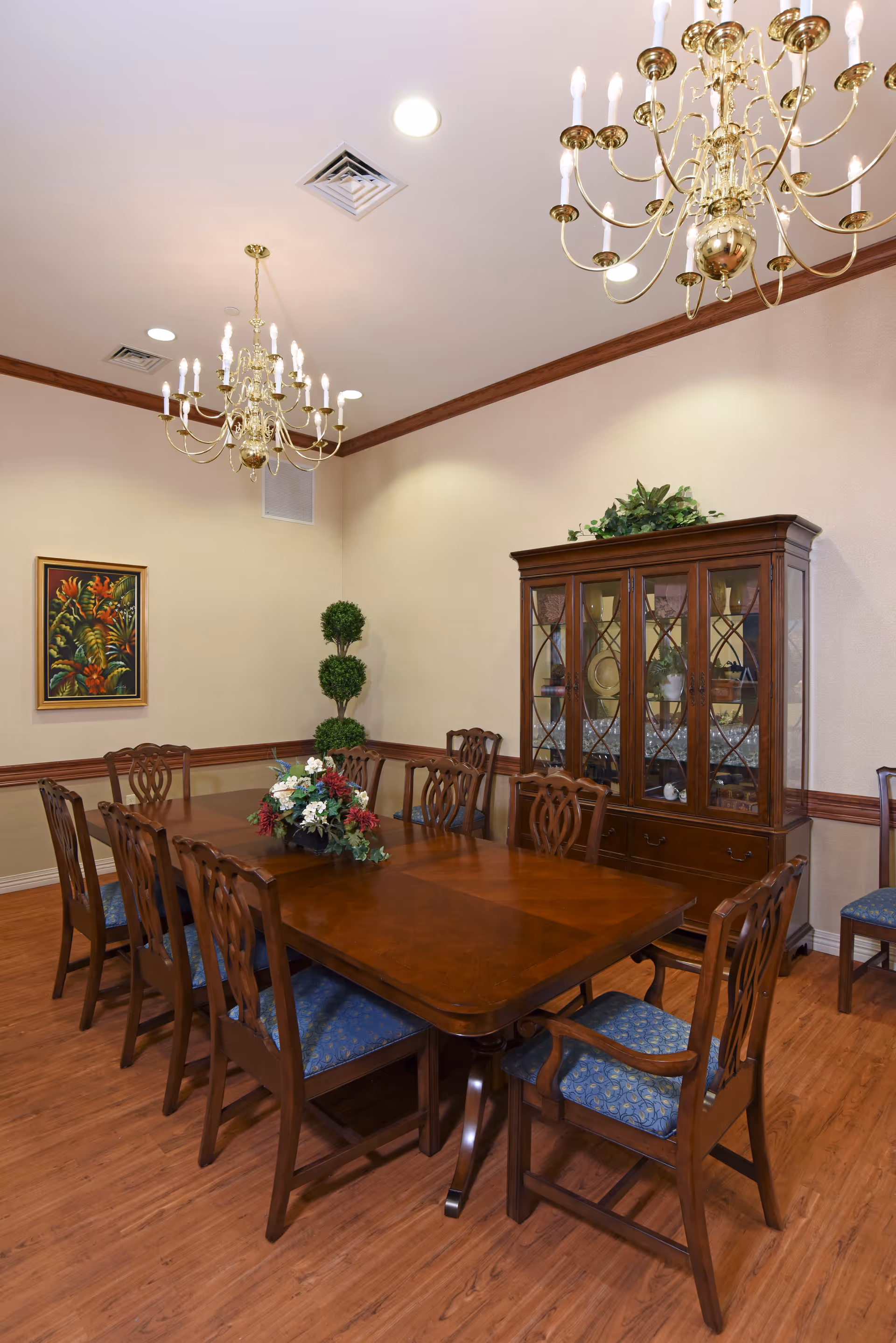 A formal dining room with a large wooden dining table surrounded by eight wooden chairs with blue cushioned seats. Two ornate gold chandeliers hang from the ceiling. A wooden china cabinet with glass doors is against the wall, displaying dishes and decorative items. A framed floral painting and a potted topiary plant are also visible in the room.