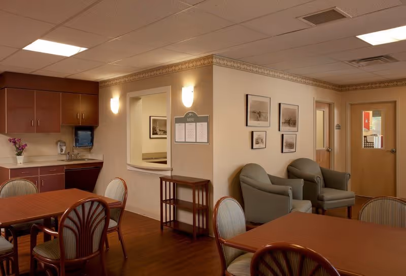 A cozy common area in a senior living facility featuring wooden tables and chairs with striped cushions, two gray armchairs against a wall adorned with framed black and white photographs, a small wooden shelving unit, and a kitchenette with cabinets, a sink, and a paper towel dispenser. The walls are beige with decorative trim, and there are ceiling lights providing illumination.