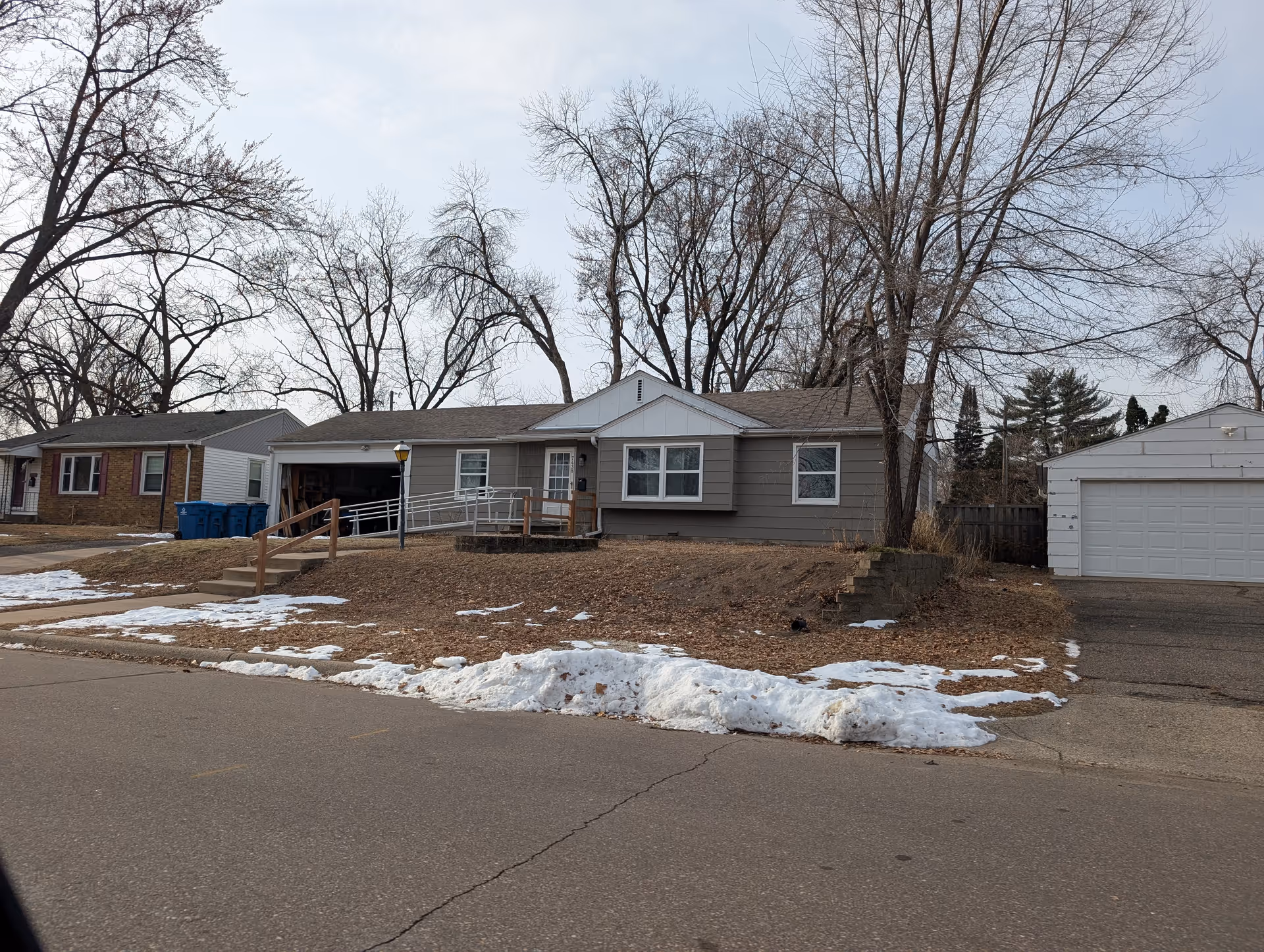 Single-story house front with a wheelchair ramp, detached garage, bare trees, and patches of snow on the yard and street.