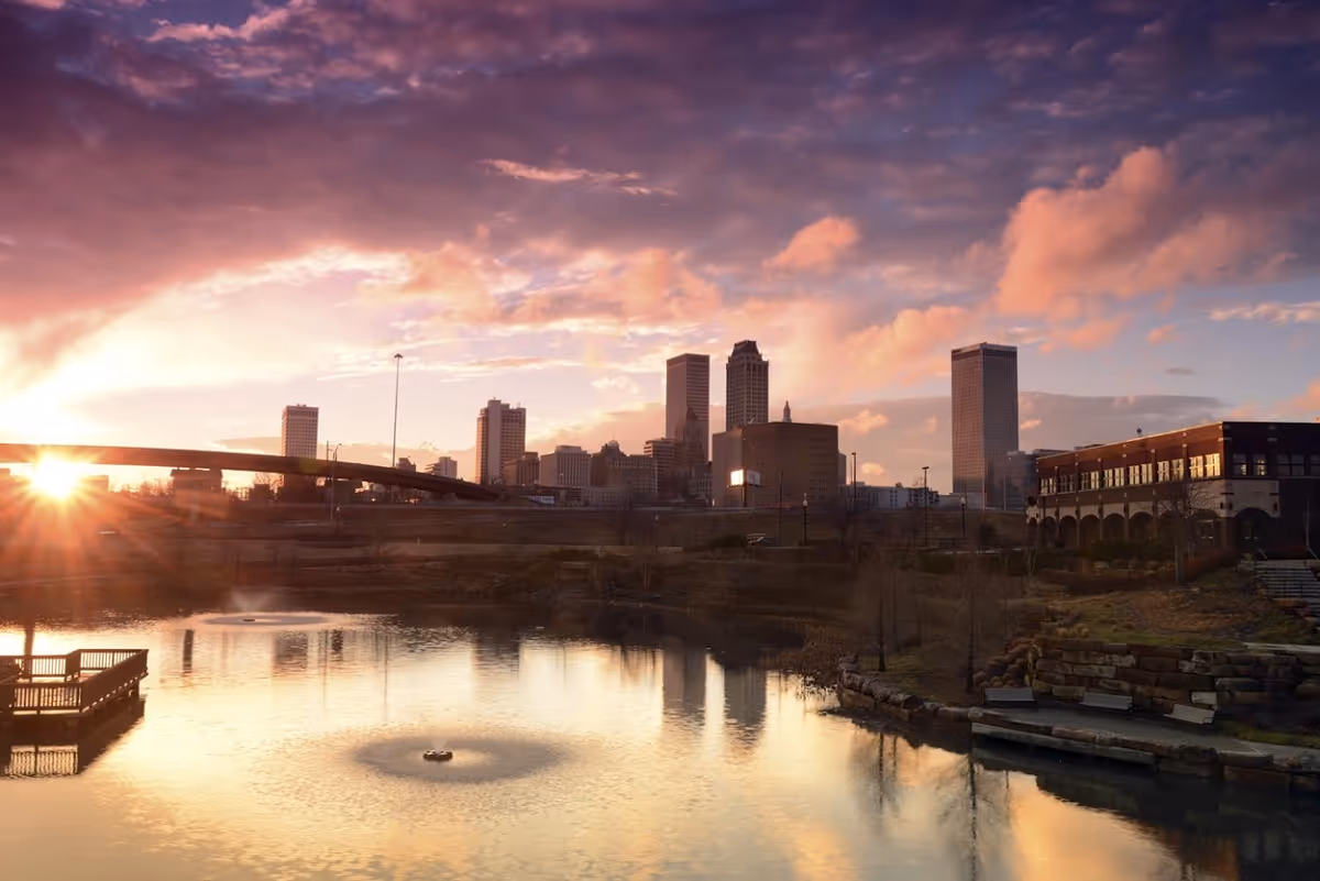 Sunset over a city skyline reflected in a calm river beside a wooden waterfront.
