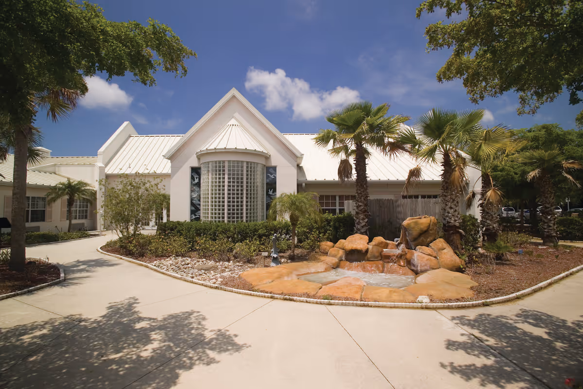 Exterior view of a single-story building with a white roof and a large glass block window in the center. The building is surrounded by landscaped greenery including palm trees, bushes, and a rock water feature in the front. The sky is clear with a few clouds.