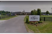 Driveway curves toward a grand entrance building with a sign and wooden fencing.