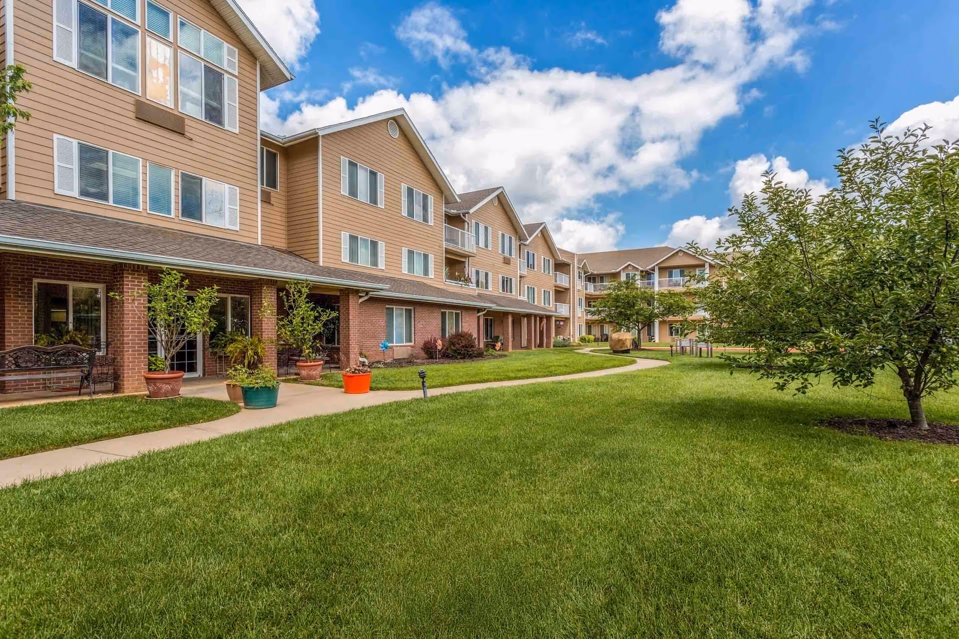 Three-story senior living building with brick and beige siding facing a green courtyard and walkway under a partly cloudy sky.