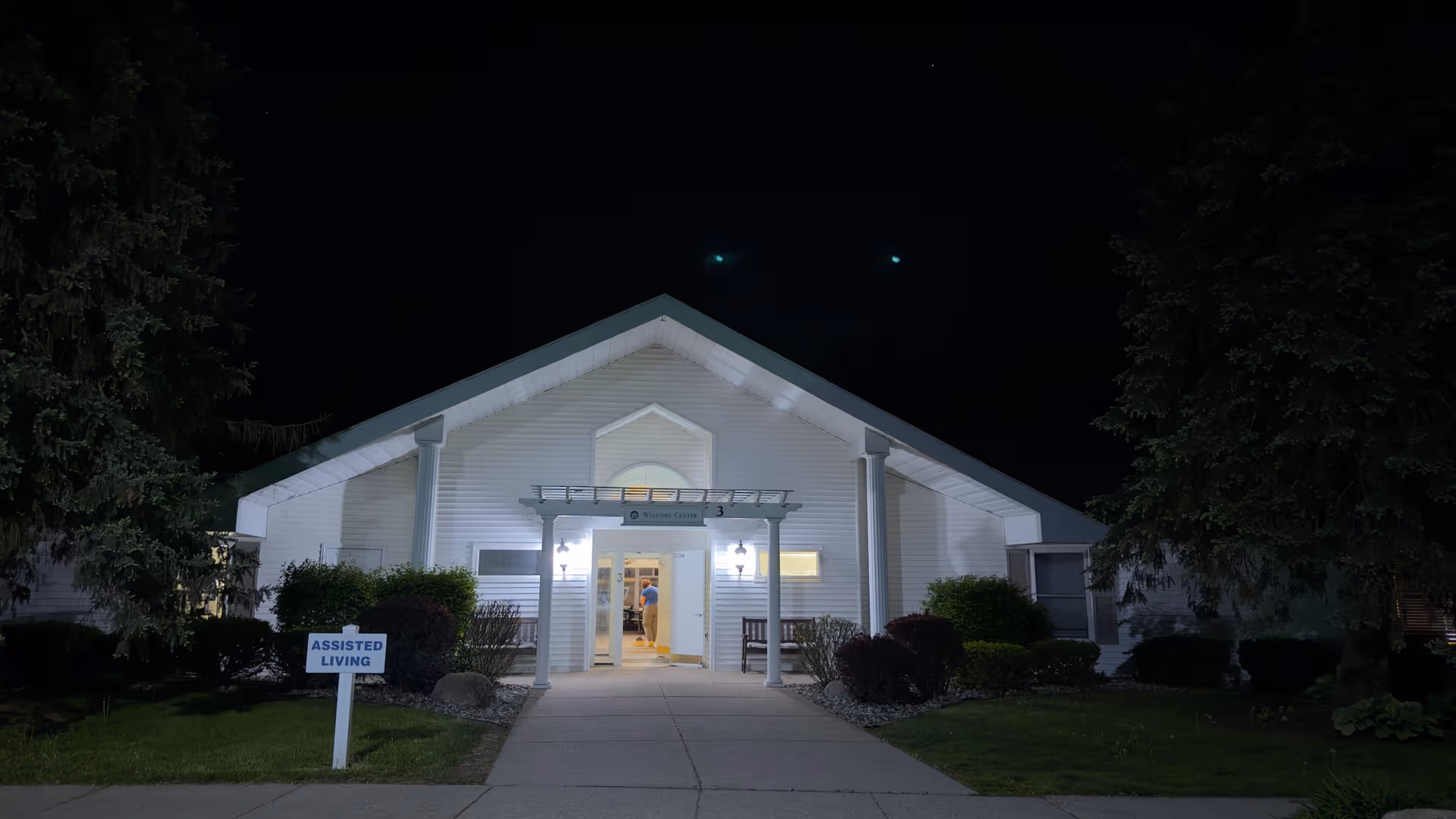 White assisted-living facility front entrance illuminated at night, shrubbery on either side.