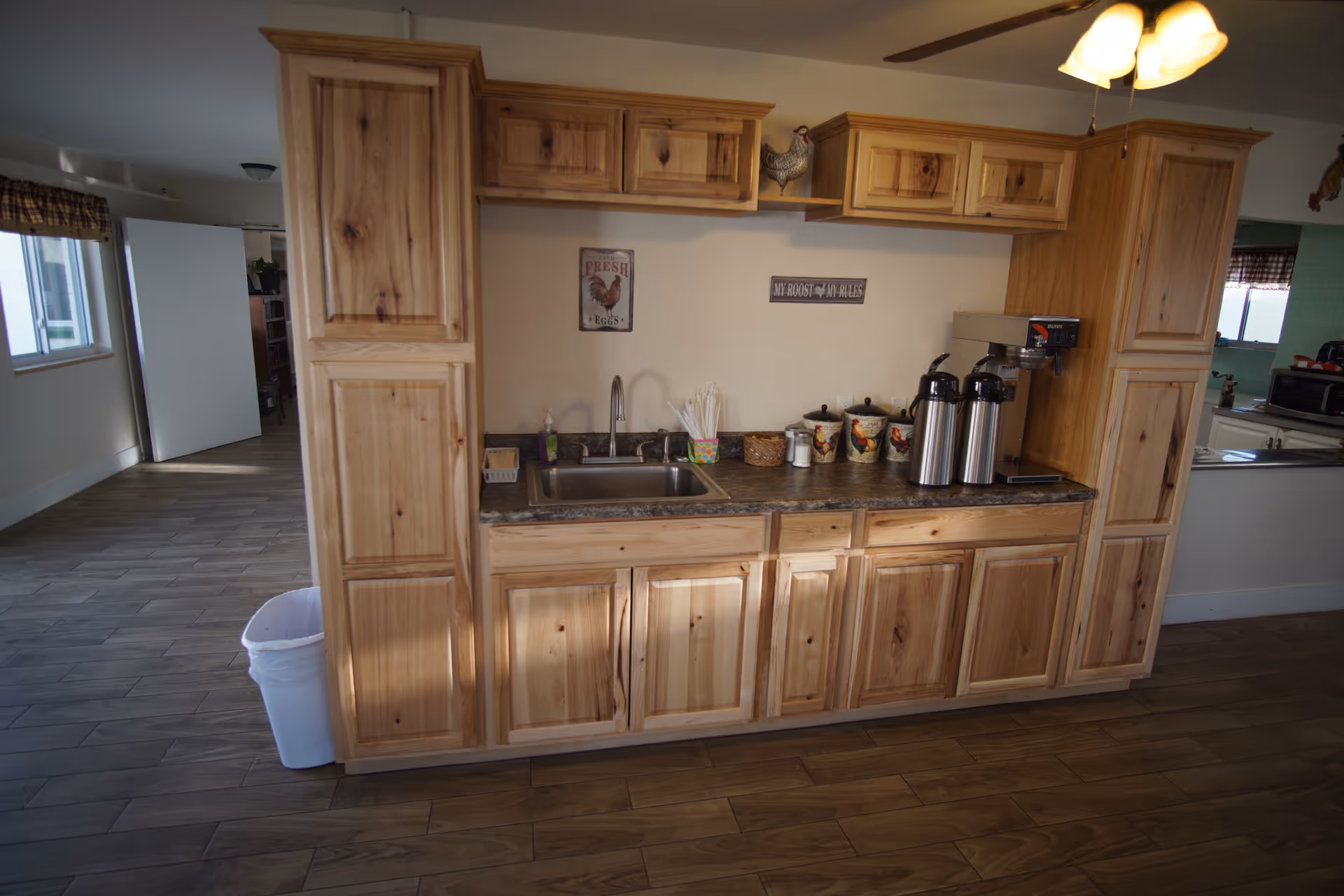 A kitchenette area with wooden cabinets and a countertop. The countertop has a sink, two stainless steel coffee dispensers, a coffee maker, and several containers. Above the sink, there are two decorative signs on the wall and a small shelf with a decorative chicken figurine. The floor is wood-patterned, and there is a ceiling fan with lights above.
