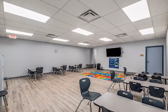 Interior activity room with tables, chairs, a colorful mat, and a mounted TV.