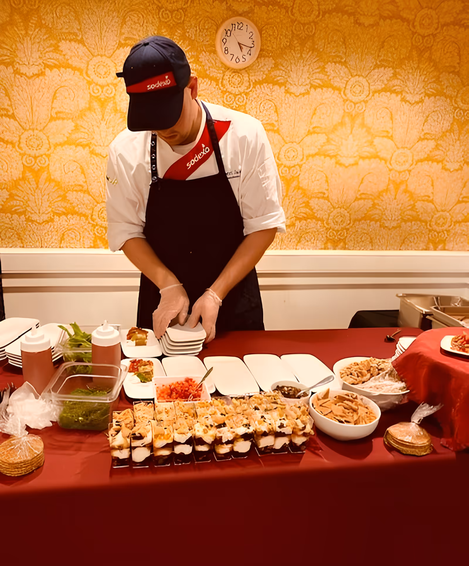A chef wearing a black cap and apron with a white shirt is preparing plates on a table covered with a red tablecloth. The table has various food items including sushi, sauces, and bowls of chips. The background features a yellow patterned wallpaper and a clock showing the time as 4:15.