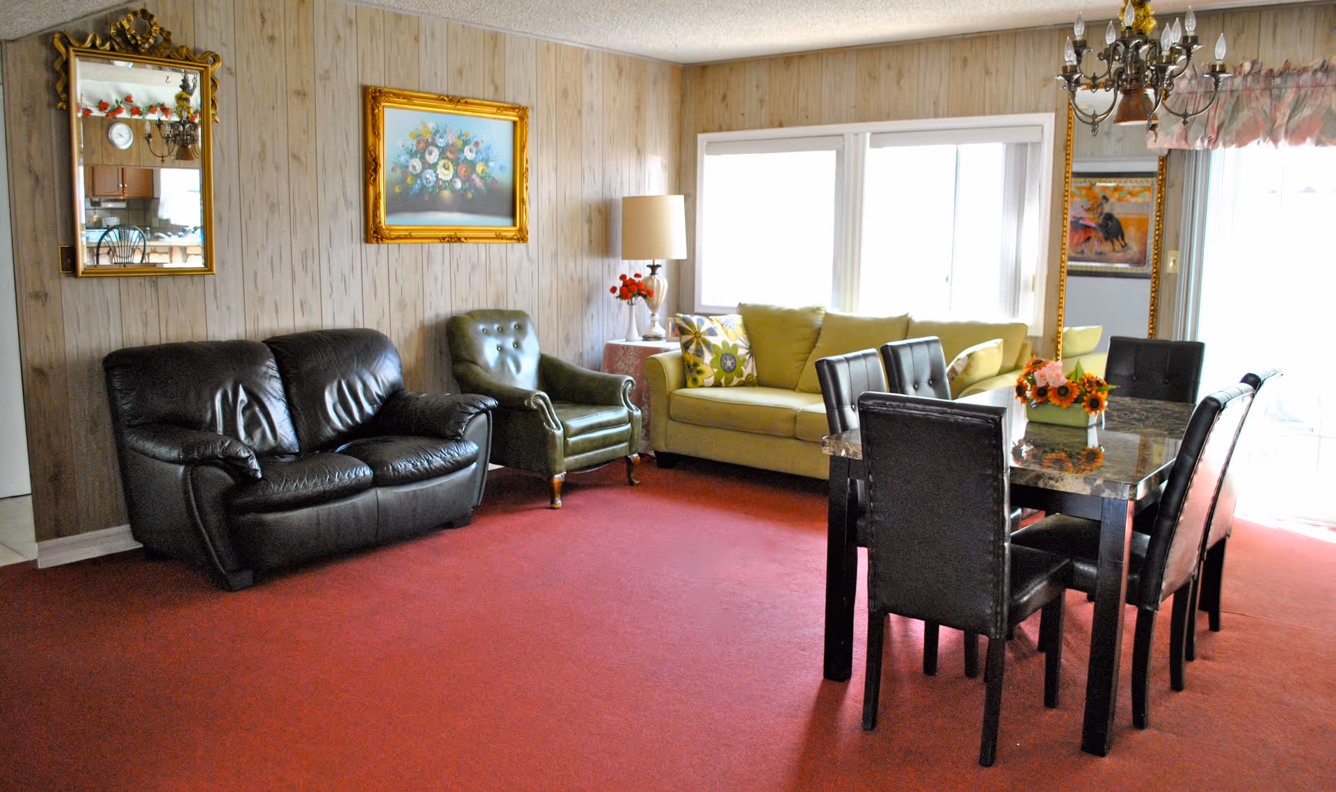 A cozy living room with a red carpet, featuring a black leather loveseat, a green armchair, and a light green sofa with patterned cushions. There is a dining table with six black chairs, a chandelier above it, and a large window letting in natural light. The walls are wood-paneled with a floral painting and a decorative mirror.