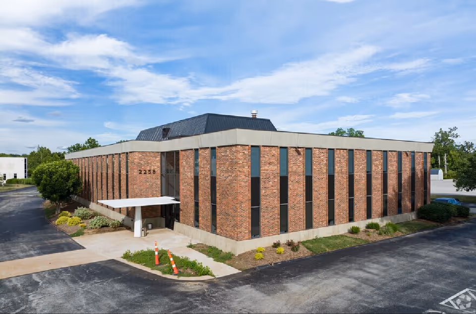 Brick building with a front entrance canopy, tall narrow windows, and a paved area.