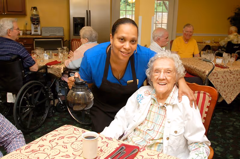 A smiling caregiver in a blue shirt and black apron is serving coffee to a happy elderly woman seated at a dining table in a senior living facility. Other elderly residents are seated at tables in the background, some in wheelchairs, in a warmly lit dining room with patterned tablecloths and large windows.