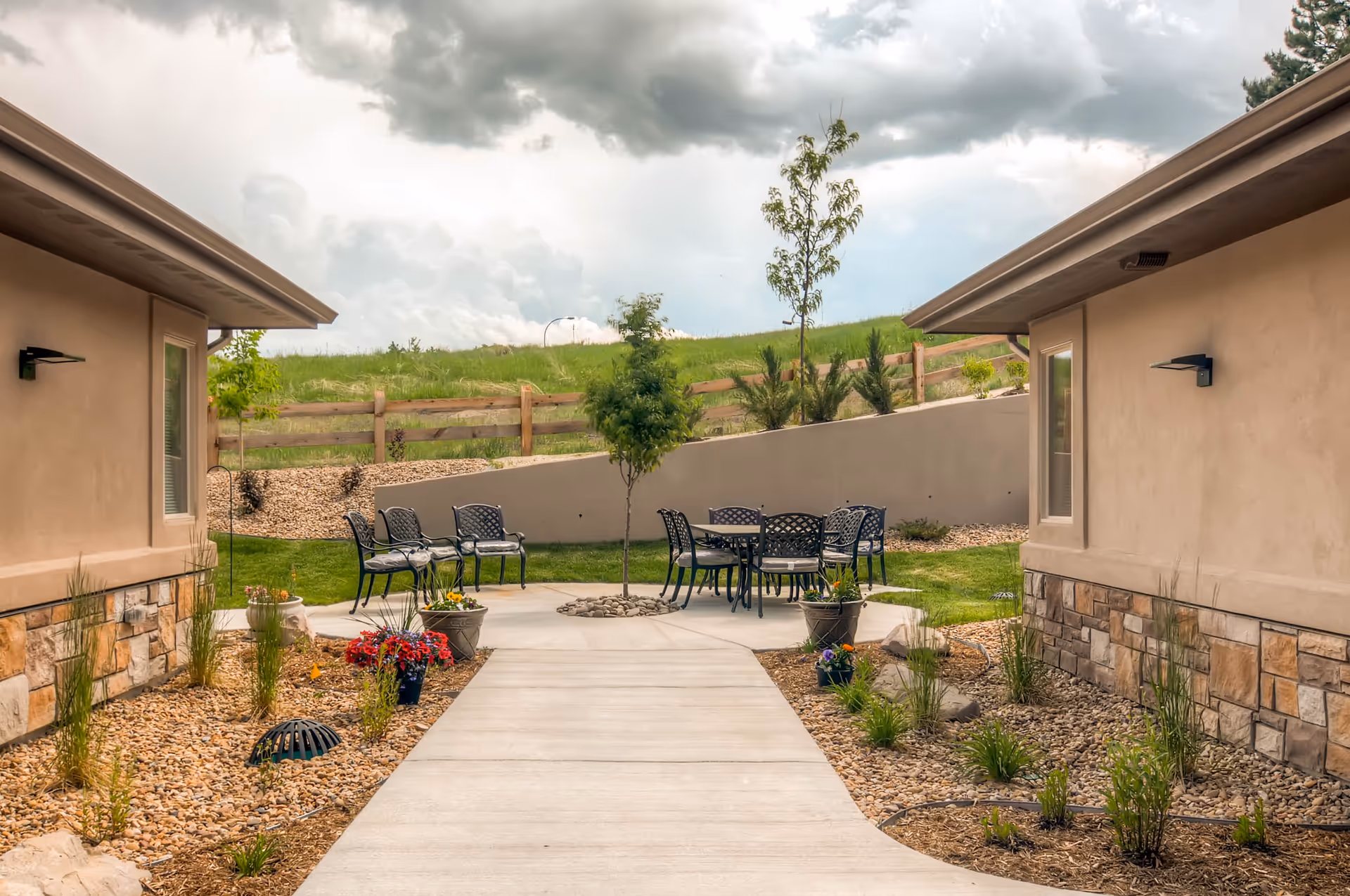 Outdoor courtyard with black metal tables and chairs between beige buildings and a landscaped path.