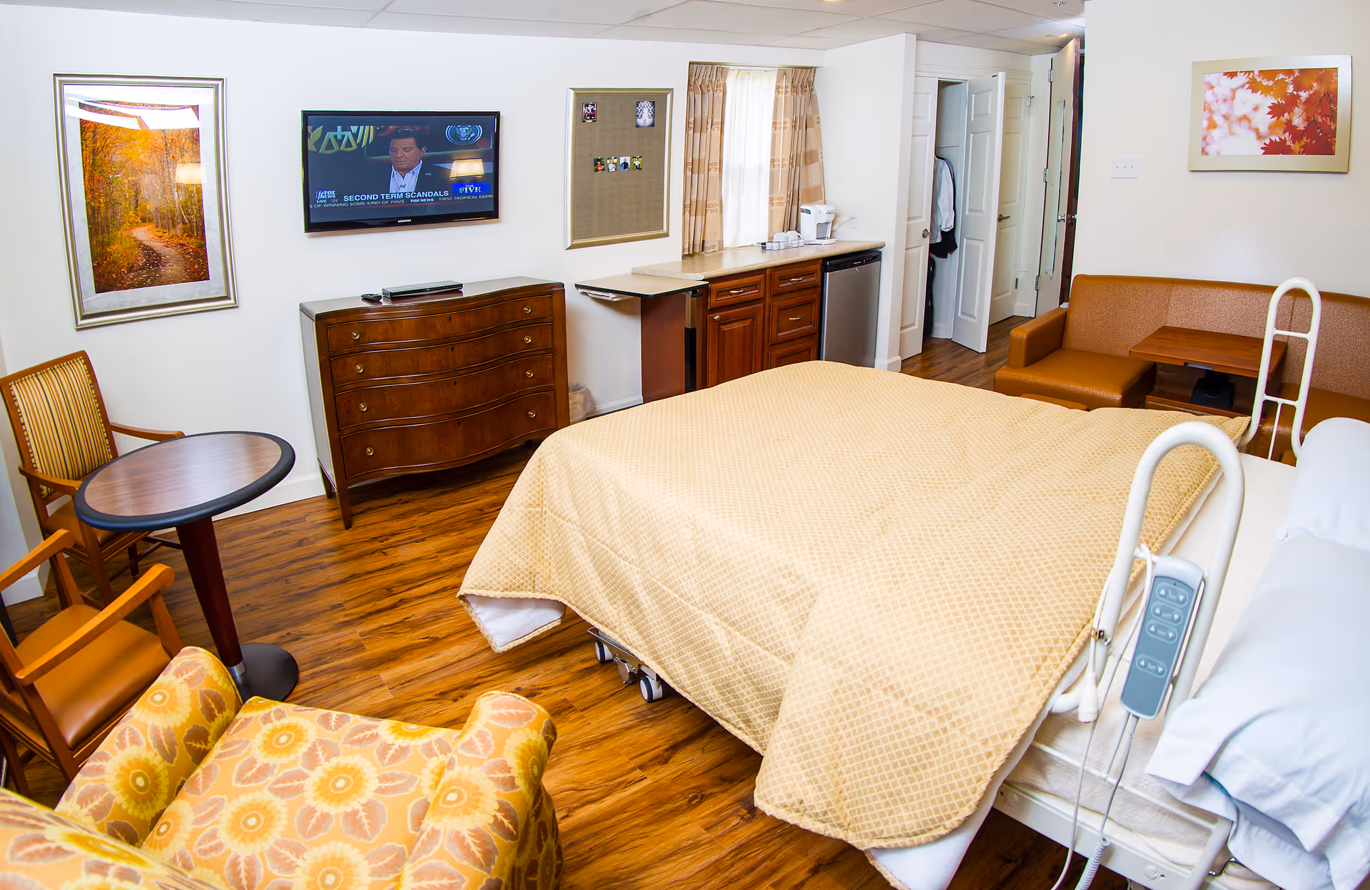 A hospital-style patient bedroom with bed, dresser, seating area, and kitchenette.