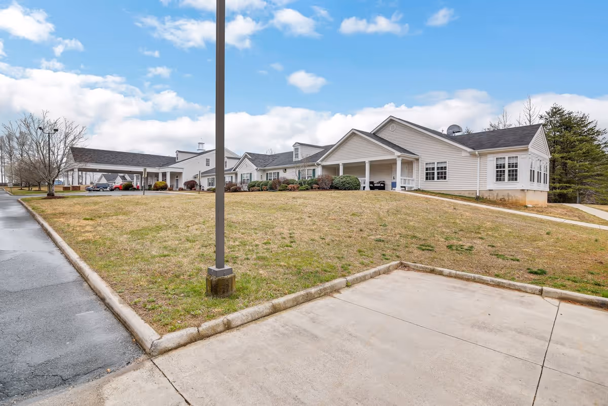 Exterior view of a single-story senior living facility building with white siding, a covered entrance, and a grassy area in front under a partly cloudy blue sky.