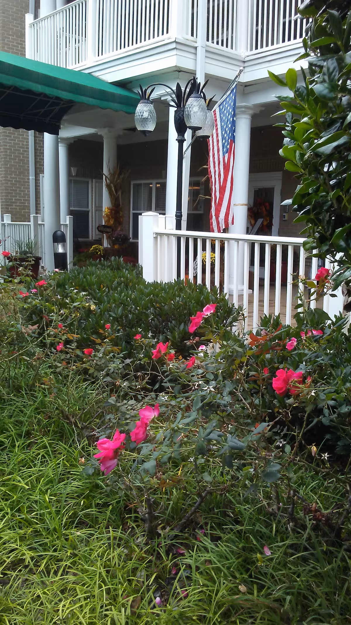 Front exterior of a senior living facility with porch, railing, flag, and flowering garden.