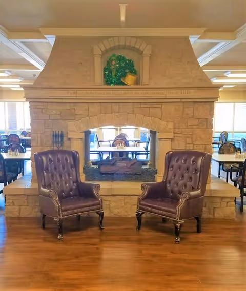 Two dark brown leather tufted armchairs positioned in front of a large stone double-sided fireplace in a spacious room with wooden floors and multiple dining tables and chairs in the background.