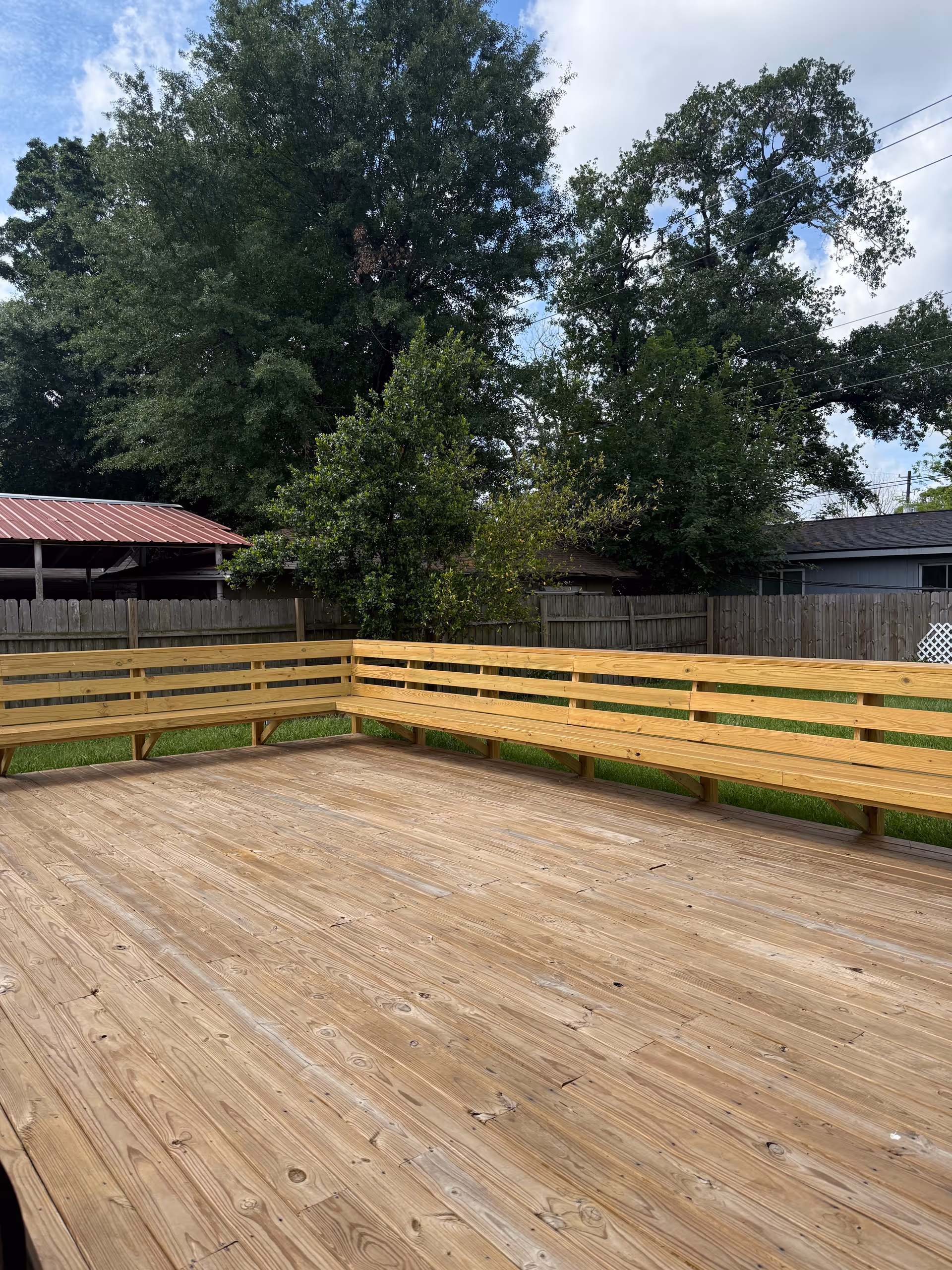A large wooden deck with built-in benches, surrounded by trees and a fence outdoors.
