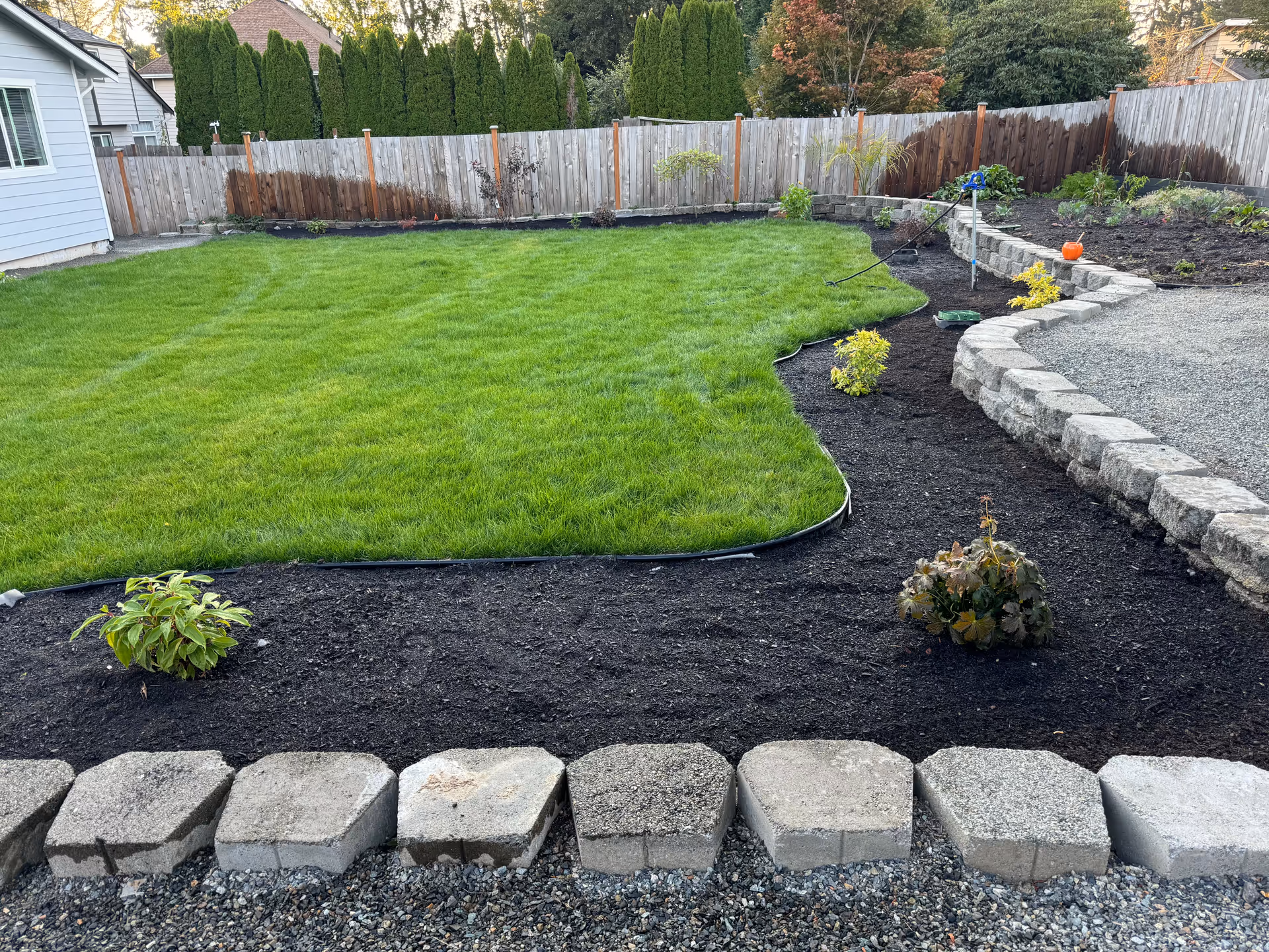 Well-maintained backyard with green lawn, stone edging, and garden beds behind a wooden fence.