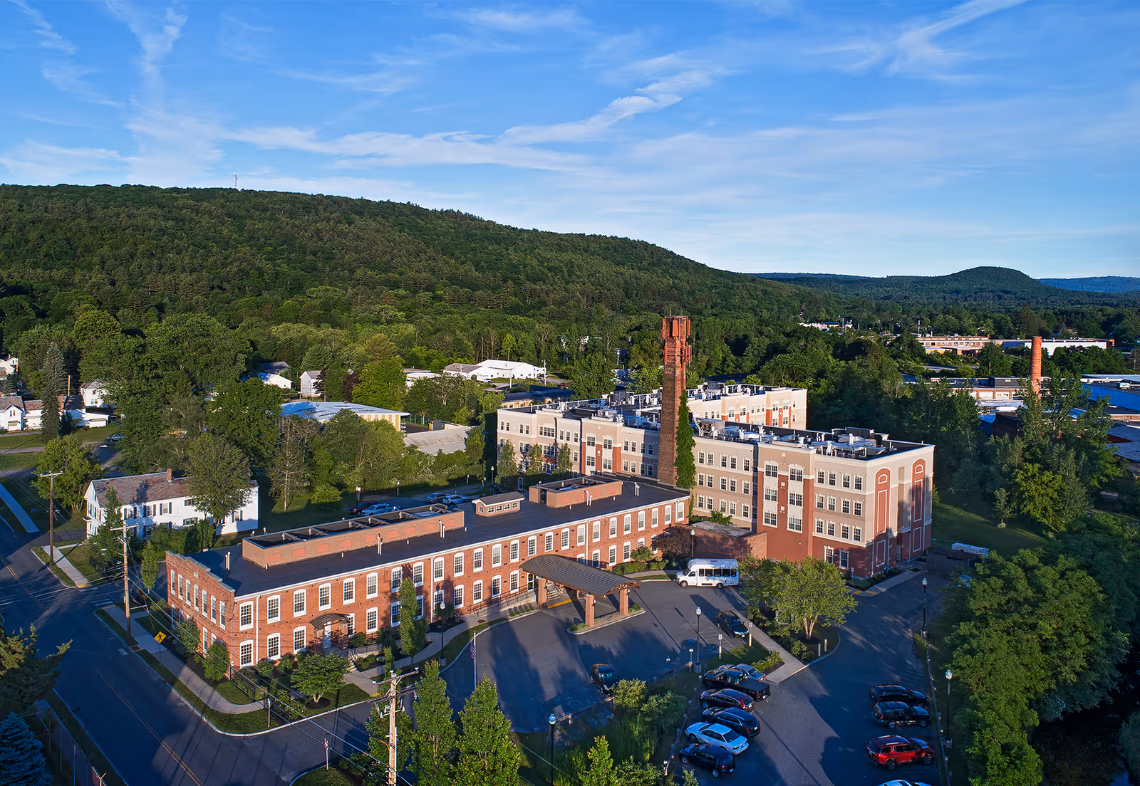 Aerial view of the American House Keene facility, showing a large multi-story brick and beige building with a tall chimney, surrounded by trees, parking lots, and nearby roads, with forested hills in the background under a blue sky.